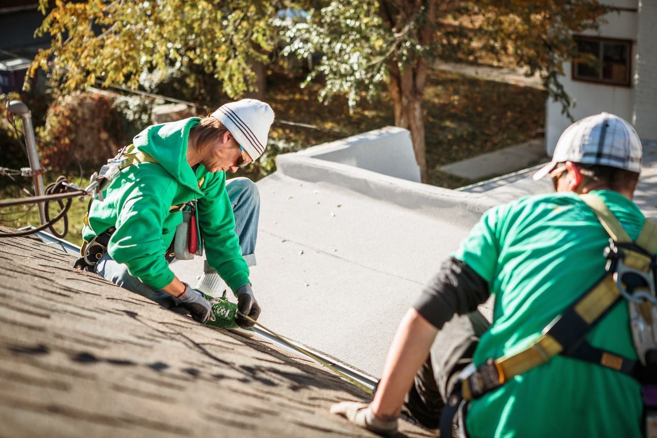 Two workers with safety harnesses repairing a shingle rooftop on a sunny day